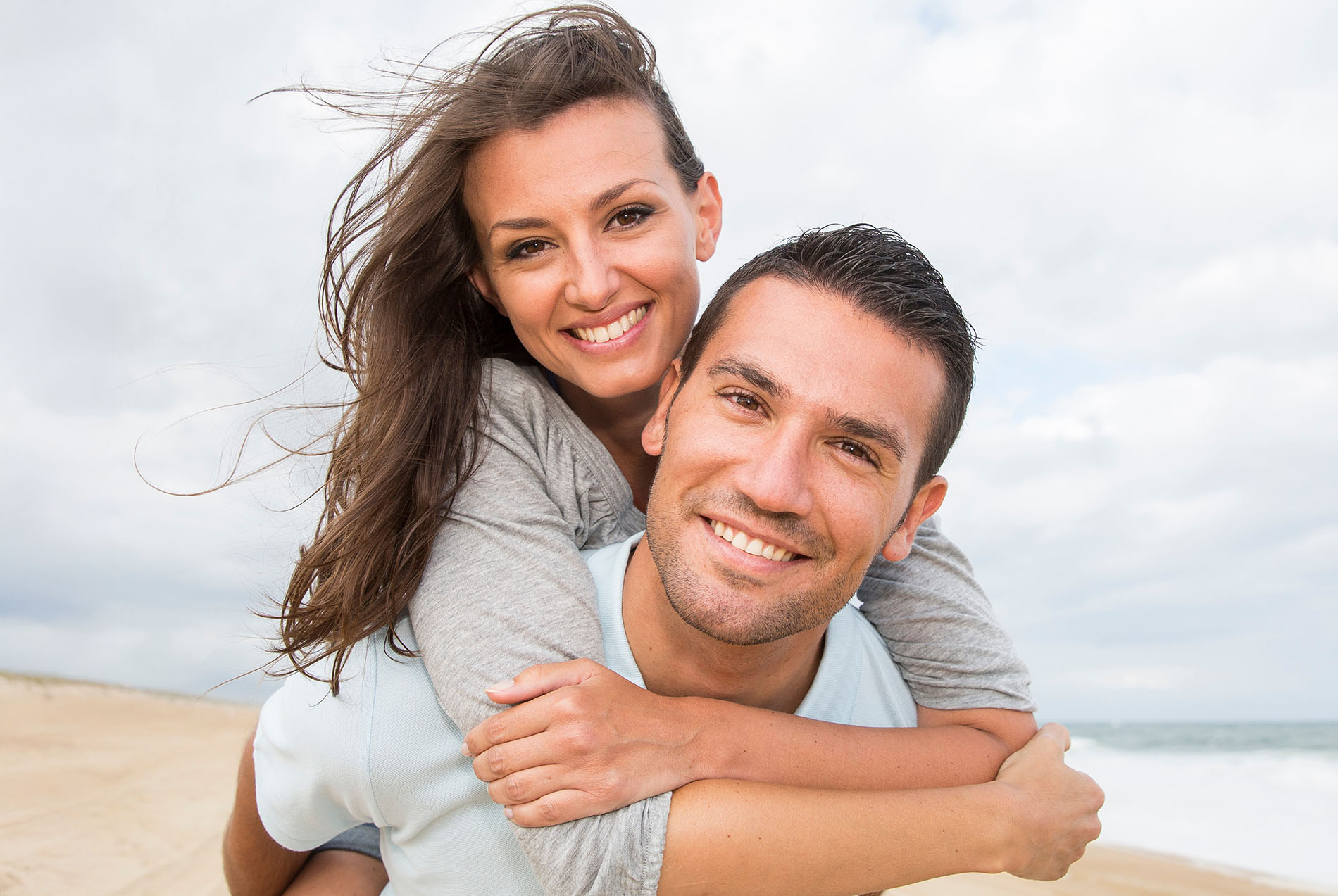 The image shows a man and woman embracing each other on a beach, with both smiling and looking at the camera.