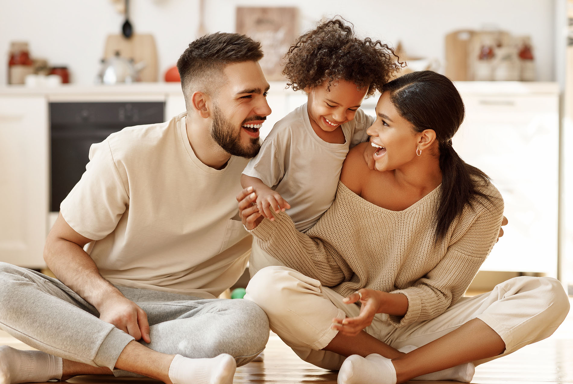A family of four poses together, smiling at the camera, with a man sitting on the floor and two children standing beside him, while a woman kneels behind them, all dressed in casual attire.