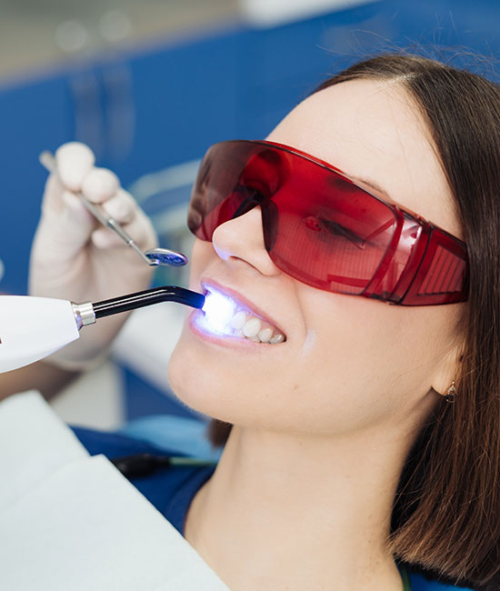 A woman wearing red goggles is sitting in a dental chair with her mouth open, receiving a dental treatment using a device that appears to be a toothbrush-like tool being applied by a professional.