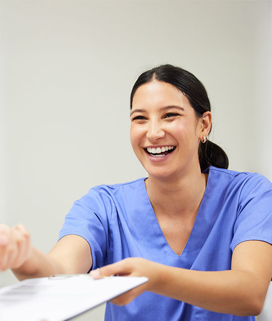 The image shows a smiling woman wearing scrubs, holding a clipboard, and handing something to another person who is out of frame.